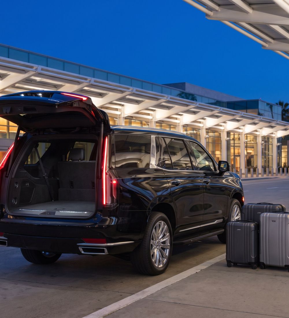 Suitcases, a golf travel bag and a folded stroller on the curb beside the open rear of the Black Cadillac Escalade